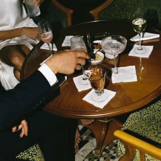 A couple in evening wear sip Bellinis on an outdoor terrace, at a table with antipasta nibbles and cocktails