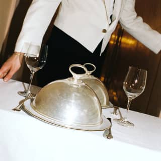 A waiter pushes a table for two, with wineglasses, cutlery and plates covered with silver cloches, in a room service detail.