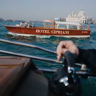 Hotel Cipriani's red boat with white canopy sails in front of Basilica Santa Maria della Salute, seen from a nearby vessel.
