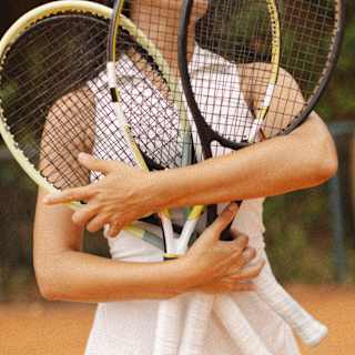A person in a white tennis kit holds three tennis racquets crossed in front of their chest on an outdoor clay court. Their face is not visible.