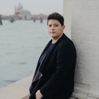 Head Chef, Vania Ghedini, with short dark hair and black jumper, leans on a wall with views across the canal to the Basilica.