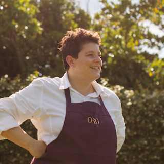 Oro Head Chef Vania Ghedini, with short dark hair, wearing a branded apron, smiles in a portrait taken in the hotel gardens.
