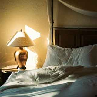 A sunlit bedroom with unmade white bedding, a wooden headboard, a canopy frame, and a bedside table with a lamp casting warm light and shadows on the beige wall.