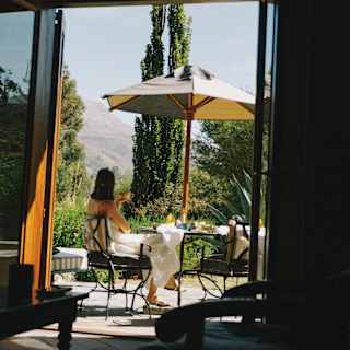 A woman sits at a terrace table with coffee, shaded by a parasol, facing the views, seen from a room interior through a door.