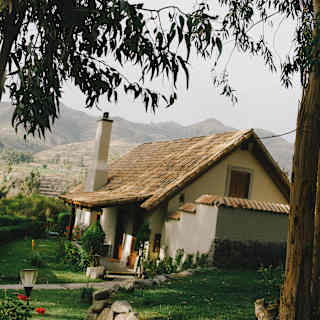 Slightly angled shot of a residence with a steep-gabled roof, tucked into the Las Casitas gardens, framed by tall trees.