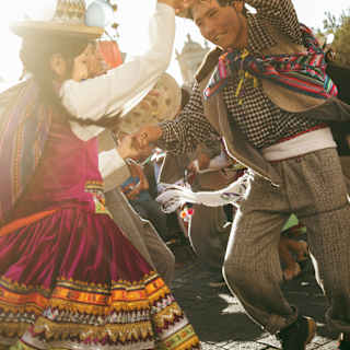 A local woman, in a cherry red dress with gold and yellow stripes, dances with a man in a checked shirt on Arequipa Day.