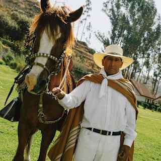 A person wearing a wide-brimmed hat, white shirt, and traditional brown-and-white poncho stands outdoors on grass holding the reins of a brown horse, with trees and hills in the background.