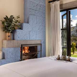 Open doors bring sublime canyon views into a spa room with a bed and a roaring fire in a lilac-painted brickwork fireplace.