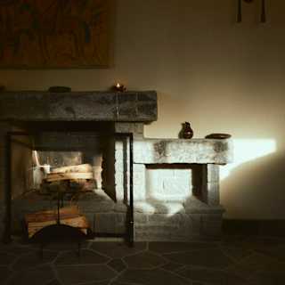 A dimly lit stone fireplace with logs stacked below, small decorative objects on the mantelpiece, a painting above, and a wall sconce with two lights casting a warm glow on the wall. Sunlight streams in from the right.