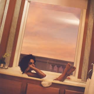 A woman with dark curly hair soaks in a foam bath with her feet up on the sill of the bathroom window, framing the sunset.