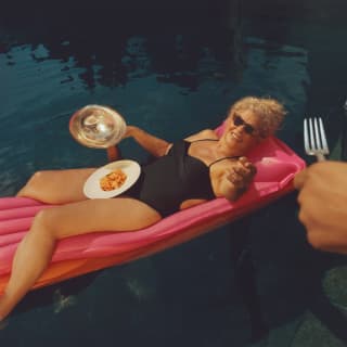 A hand passes a fork to a blond guest in a black bathing costume, floating on a pool lilo with a bowl of pasta in her lap.