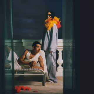 On a balcony at night, a woman poses in grey pyjamas, feather boa and shades by a man in white casuals reclining on a bench.