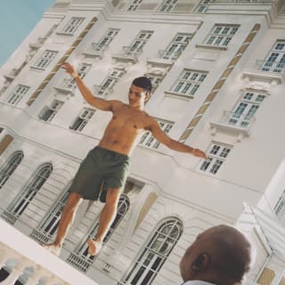 A male staff member watches as a topless young man in green shorts balances on the white balustrade wall outside the hotel.
