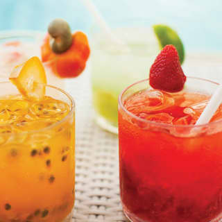 Bright coloured cocktails sit poolside in elegant tumblers, including an orange maté with passionfruit and a red fruit caipirinha