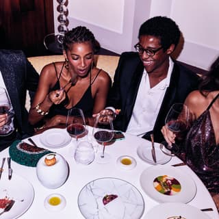 Five young people in party dress sit at a round table laughing and drinking red wine while sharing plates of dessert