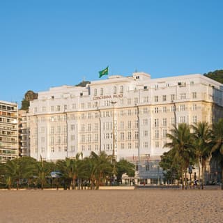 A giant Brazilian flag flies from the roof of the art deco Copacabana Palace Hotel. In the foreground palm trees and beach