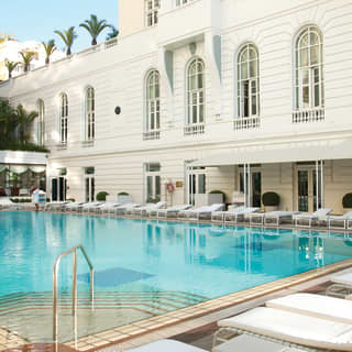 Outdoor restaurant pool surrounded by sunbeds in front of a white hotel facade