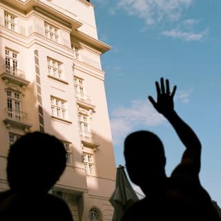 Behind the silhouette of two guests, a corner façade of the Copacabana Palace rises against the blue sky in soft-white stone.