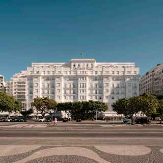 View of the imposing, grand white Copacabana Palace edifice as it rises against a blue sky behind a row of mature trees.