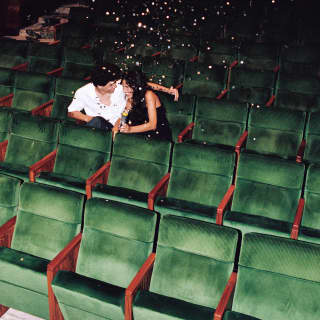 With confetti in the air, a man in a white shirt and a woman in black cosy up in the green velvet seats of the empty theatre.
