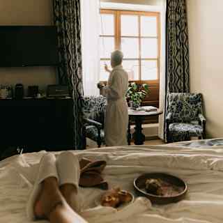 A guest stretches her legs on a bed alongside plates of pastries, as a man in a robe adjusts drapes to let in morning light.