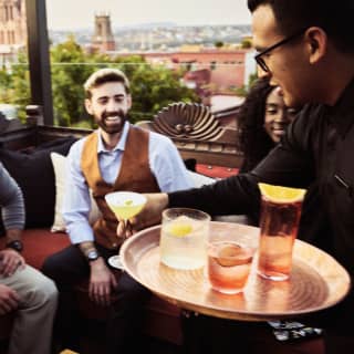 A smiling group are served cocktails on sofas at the Tunki bar with rooftop views of the historic city behind