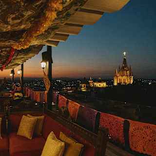 A spot-lit parroquia with an illuminated cross glows in the dark cityscape, in a moody red sunset view from the rooftop bar.