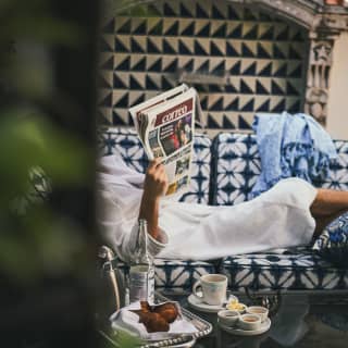 Partly obscured, a guest in a bathrobe reads a newspaper lying on a sofa next to a coffee table with a continental breakfast.
