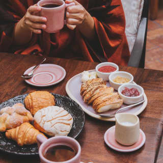 In Andanza, a woman holds a pink coffee cup at a breakfast table with traditional breads, concha buns and pain au chocolat.