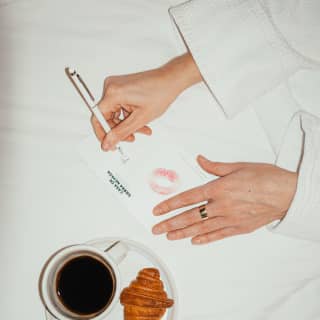 Lady writing a note on her bed with a black coffee and a croissant