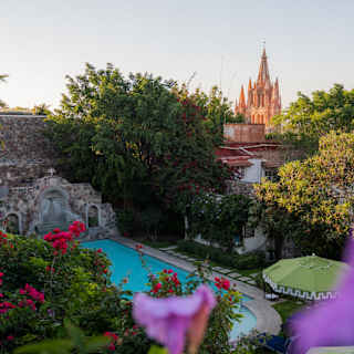 View over the shady hotel pool nestled in lush gardens to the Parroquia San Miguel Arcángel behind, bright pink in the sun.