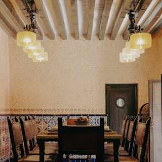 Eye-level view of a dining table and eight chairs, seen from the head, below a beamed ceiling with eight cylindrical lights.