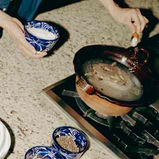 A Sazón Cooking School student adds salt from a blue bowl to a broth cooking on the hob, with other ingredient bowls in view.