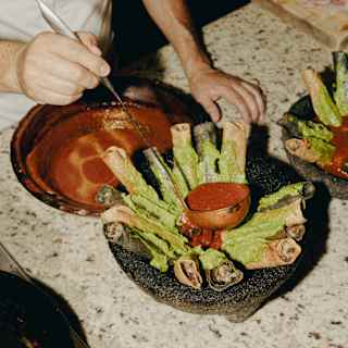 At a marble table top, a chef ladles a tomato sauce into the centre of a stone bowl containing flautas coated in guacamole.