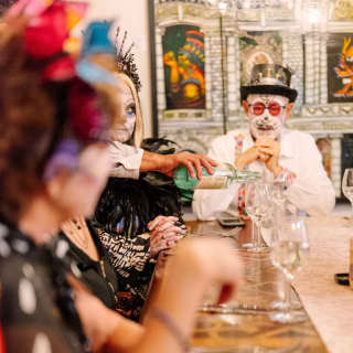 A man in pink specs, face paint and top hat sits at the end of a table of diners pouring cava, dressed for Dia de Muertos.
