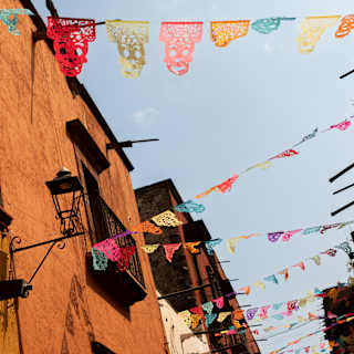 Strings of bright papel picado zigzag between the orange-rendered buildings of a San Miguel street, seen from below.