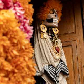 A Dia de Muertos skull-head figure stands in a doorway in large earrings and a necklace, part-obscured by a marigold garland.