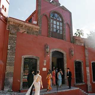 A groom and a bride, trailing a lace train, walk towards guests waiting on the frontage of the terracotta-hued Casa Parque.