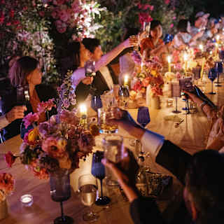 Candles glow among bright floral centrepieces in a view along a beautiful evening party table, where guests raise glasses.