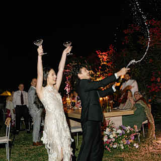A bride in an ivory slip dress raises two glasses as the groom releases a jet of Champagne from a bottle at an evening party.