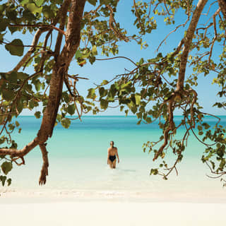 View through tree branches of a woman wading into the sea from a white sand beach in Mu Ko Ang Thong National Marine Park.