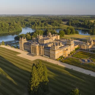 Aerial view of Oxfordshire's Blenheim Palace mansion, resplendent in sunlight, in a vast estate with lawns, lake and gardens.