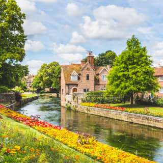 The River Stour flows past the historic, half-timbered houses of Canterbury, bordered by lawn banks of red and gold flowers.