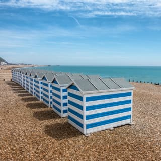 A row of twelve blue and white-striped beach huts sits on the pebble beach at Hastings, a few steps from the luminous sea.