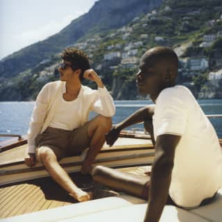 Two men in shorts and white tops, one with sunglasses, relax on a gozzo leisure boat, absorbing the hazy heat and views.