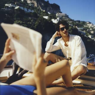 In soft foreground, a woman reads lying down while a man behind, in focus, sits and daydreams as they drift on a gozzo boat.