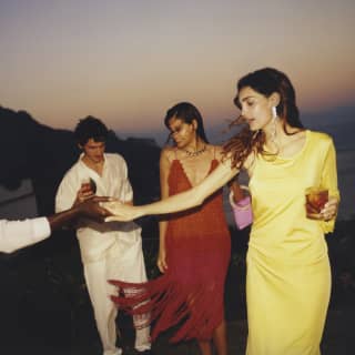 Two women, in yellow and red evening dresses, hold cocktails and dance with two men as the sky darkens at Caruso hotel.