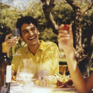 A laughing, dark-haired man in a bright yellow shirt holds a glass of white wine at a sunny lunch table with friends.