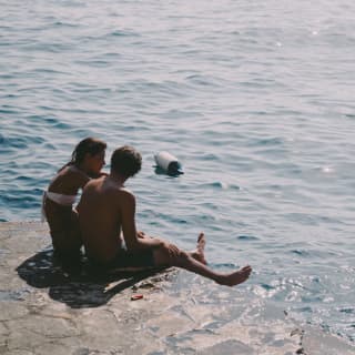 Man and woman in swimwear sitting together on a rock overlooking a harbour