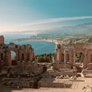 View from the Greek Temple ruins in Taormina down to the azure Ionian coast and up to the smoking, snowy peak of Mount Etna.